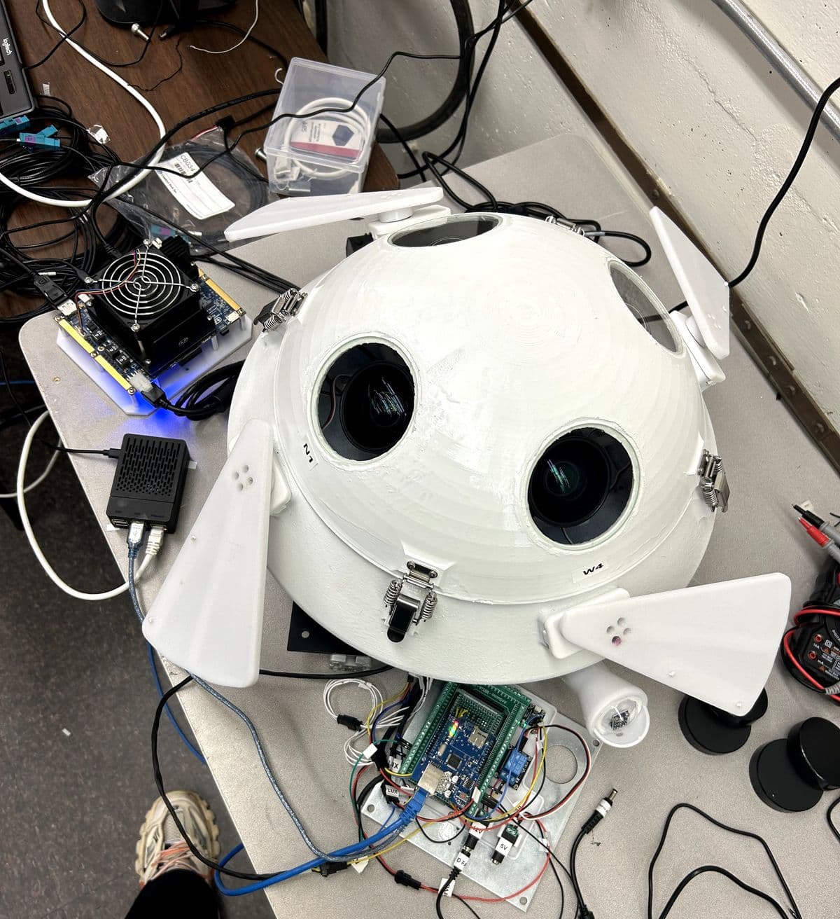 A white dome-shaped enclosure on a lab bench with four camera windows, two labeled N1 and W4, and triangular shades on hinges. Beside it are an Arduino Mega with a green screw-terminal shield and a relay, a Raspberry Pi in a black case connected to it by USB, a fan-cooled single-board computer, and a multimeter.